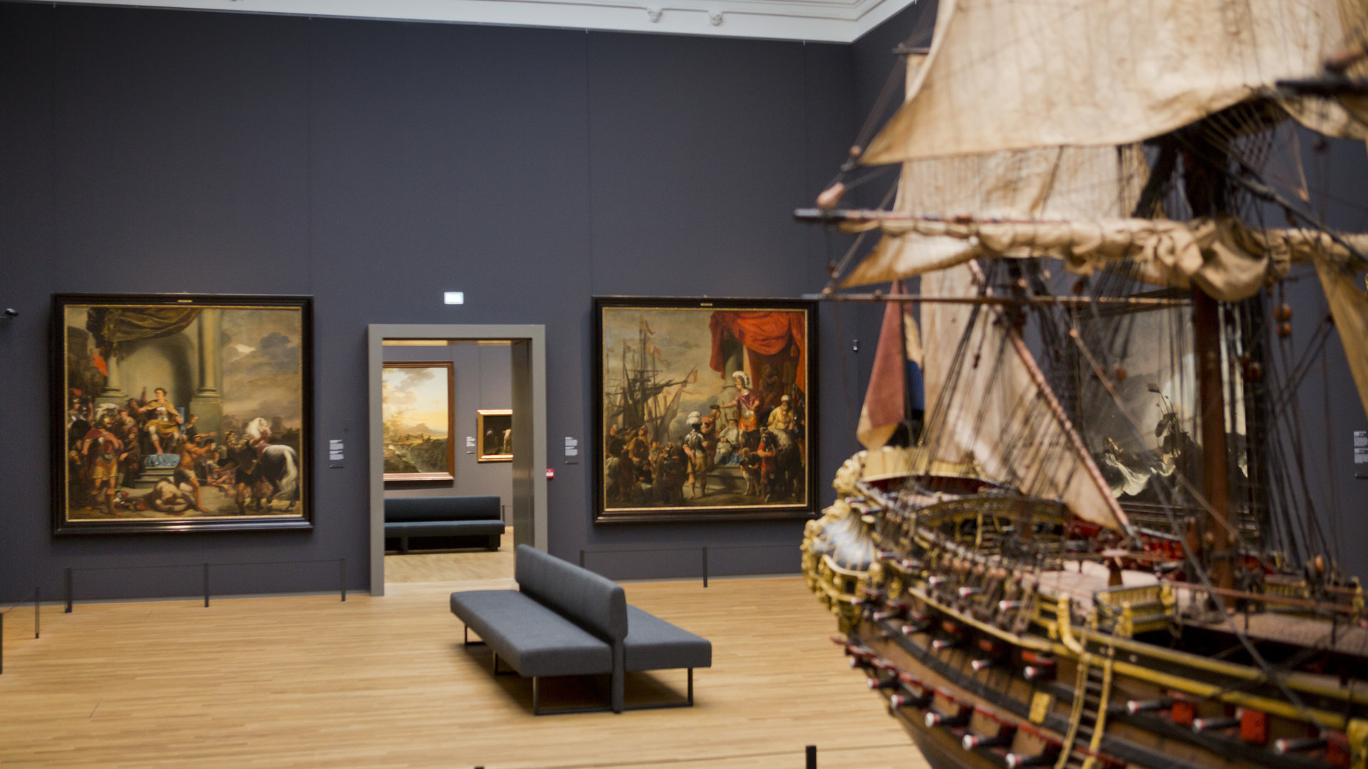 Een foto van een museumzaal waar een scheepsmodel en twee schilderijen op te zien zijn